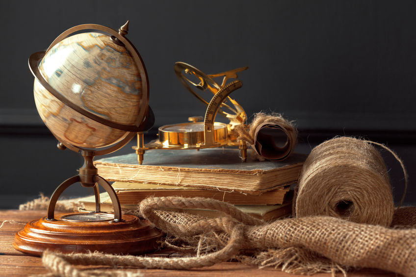 A small globe stands on a wooden desk, accompanied by old books, a brass sundial, a rolled parchment, and coiled twine, suggesting a vintage, scholarly setting.