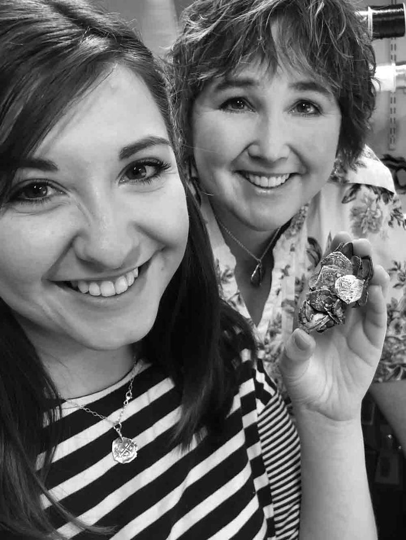 Two women smiling at the camera; one holds a clump of fused shipwreck treasure coins. They are indoors, possibly near shelves with thread spools. Both wear treasure coin necklaces.