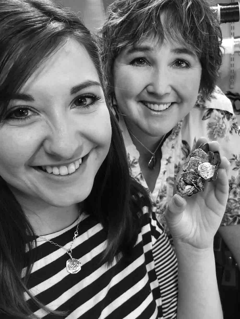 Two women smiling at the camera; one holds a clump of fused shipwreck treasure coins. They are indoors, possibly near shelves with thread spools. Both wear treasure coin necklaces.