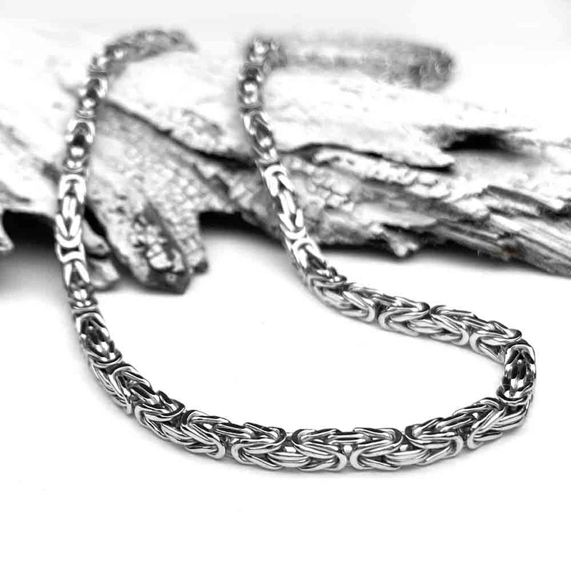 A polished silver chain lies elegantly on textured, overlapping pieces of wood, contrasting the smooth, intricate links with the rough bark-like surface.