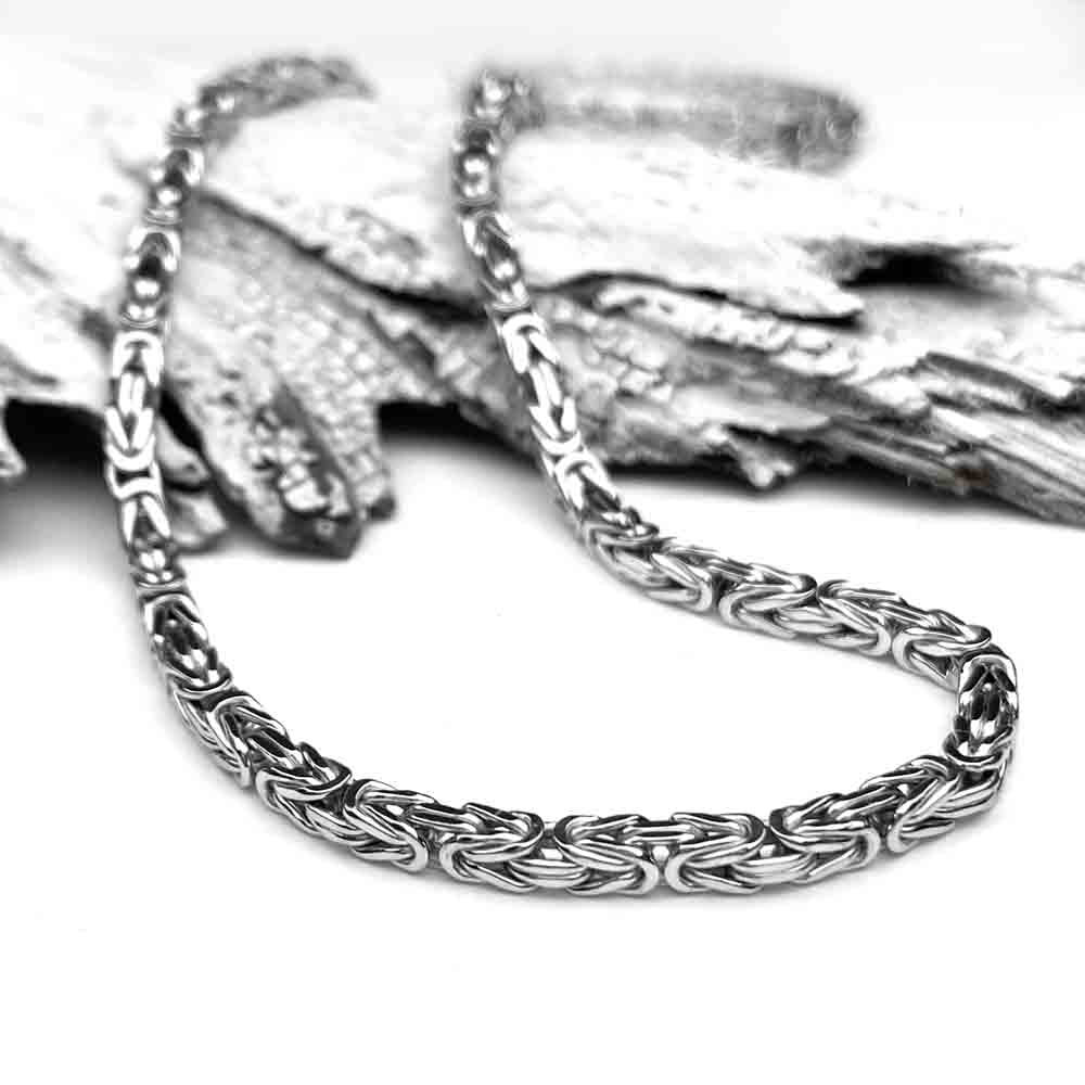 A polished silver chain lies elegantly on textured, overlapping pieces of wood, contrasting the smooth, intricate links with the rough bark-like surface.