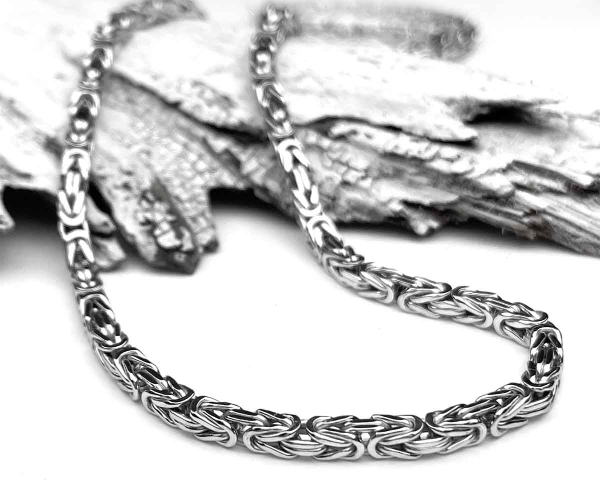 A polished silver chain lies elegantly on textured, overlapping pieces of wood, contrasting the smooth, intricate links with the rough bark-like surface.