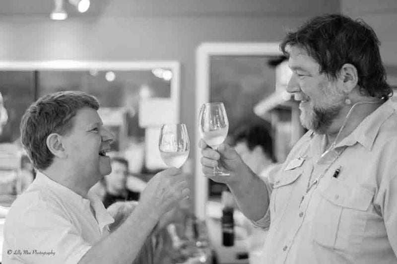 Two people clink wine glasses while smiling warmly in a lively indoor setting. A label reads "© Lilly Mae Photography."