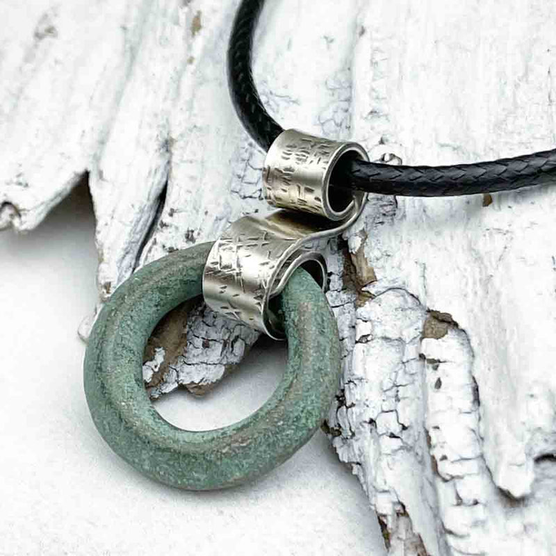 Ancient Bronze Celtic Ring Money is suspended on a hand-hammered silver bail and leather cord necklace and rests against textured white driftwood.