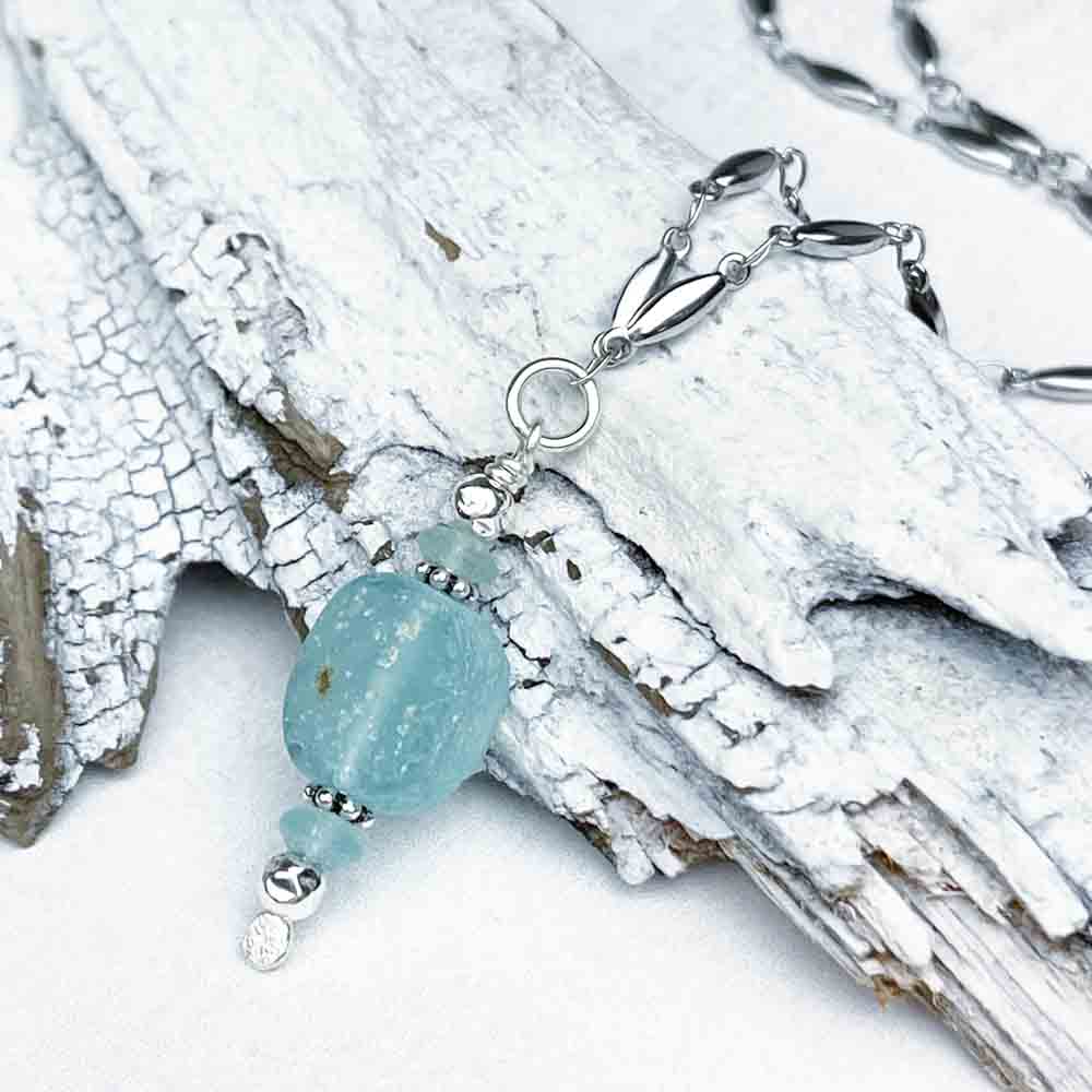 A silver and ancient Roman Glass necklace rests on textured white driftwood, highlighting the contrast between the jewelry's polished metal and the rugged wood.