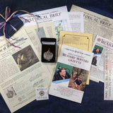 A shipwreck treasure coin pendant displayed in a black box amidst a package of documents. Text includes: "Cannon Beach Treasure Company," "Certificate of Authenticity," "Historical Brief," "Treasure Hunters Gazette," and "Coin Classification." Context is a deep blue textured surface.