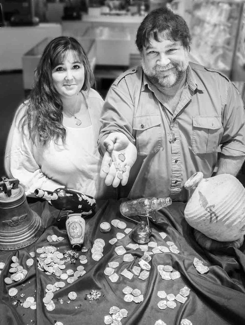 Robert and April Knecht of Cannon Beach Treasure Company are pictured smiling, with the Robert holding coins, stand behind a table displaying various artifacts, including a bell, bottles, and numerous scattered treasure coins on a dark cloth.