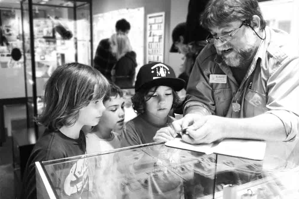 Robert Lewis Knecht of Cannon Beach Treasure Company explains treasure coins to three children at a display case in a museum. The children watch intently, surrounded by glass exhibits and visitors in the background.