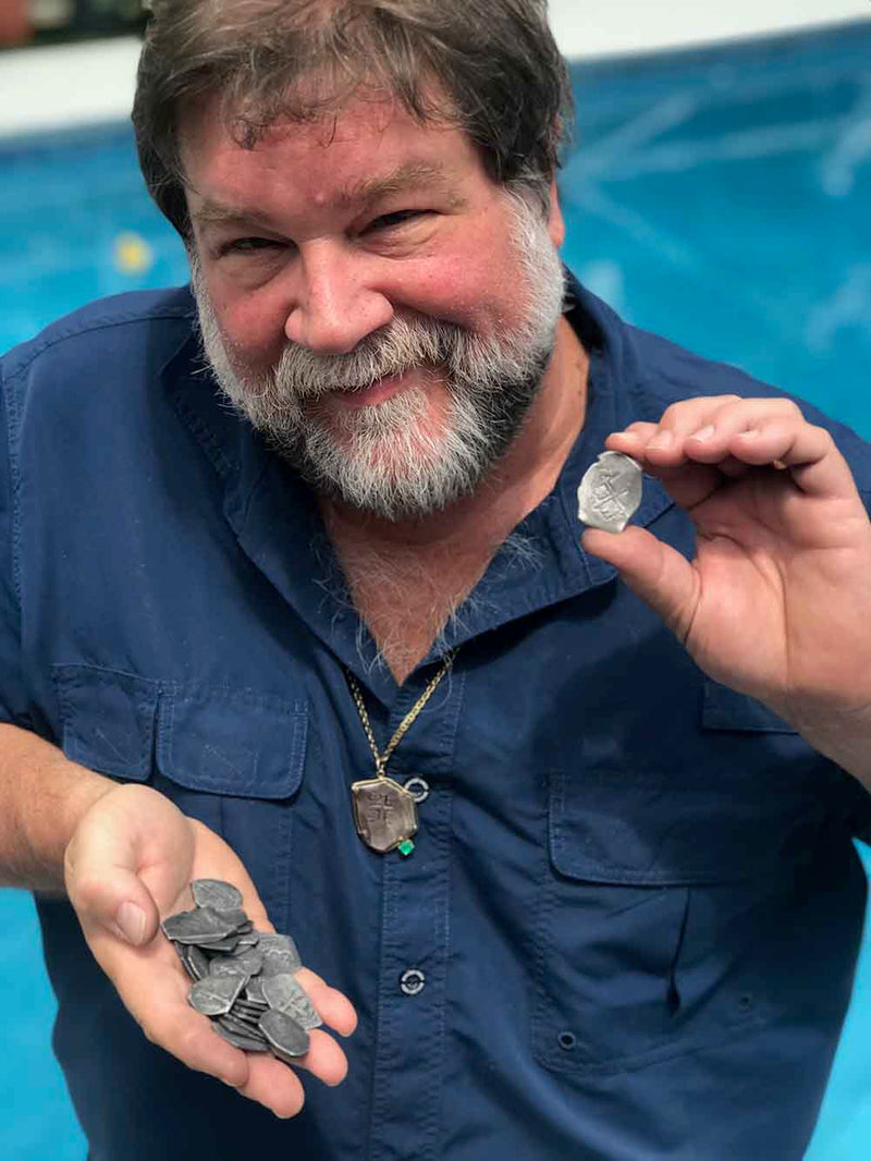 Robert Lewis Knecht holds a stack of 1715 Fleet shipwreck treasure coins in one hand and displays a single coin with the other, standing by a swimming pool, wearing a blue shirt and 1715 Fleet shipwreck treasure coin necklace.